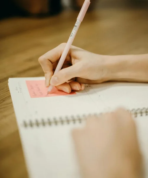 Close-up of a person writing in a notebook with a pink pen on a wooden desk.